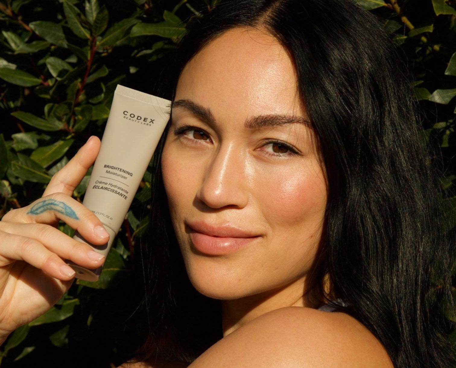 Steph Shepard holding a bottle of Codex Beauty Labs Antu Brightening Moisturizer. She's also touching the bottle to her face.